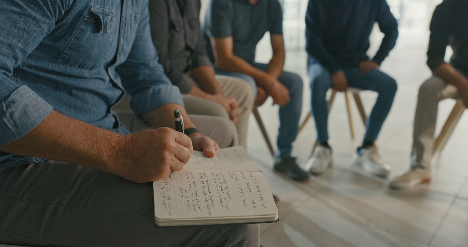 Man in group therapy writing in a notebook. Male therapist  making notes on mental health behavioral progress and key talking points during a team building activity in an interactive support session